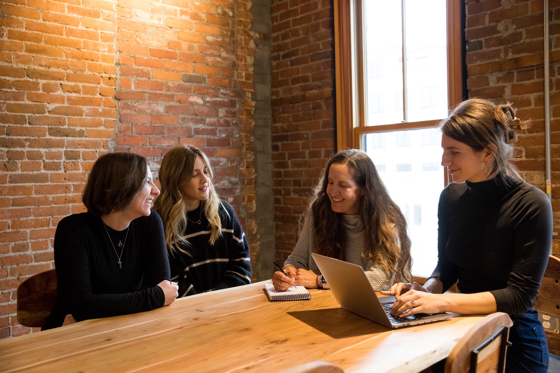 Four women engaged in a discussion around a wooden table with a laptop and notebook